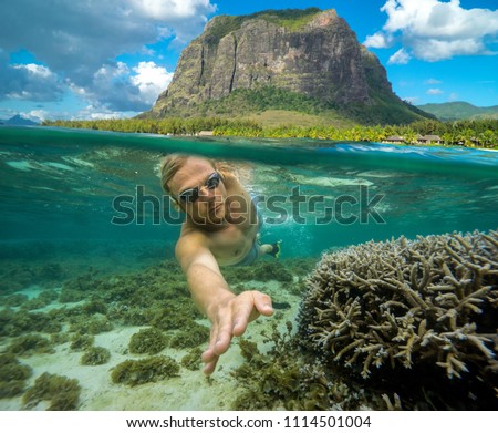 A young man dives among the multicolored corals in the Indian Ocean. Snorkeling in Mauritius. At the top of the picture there is a high mountain