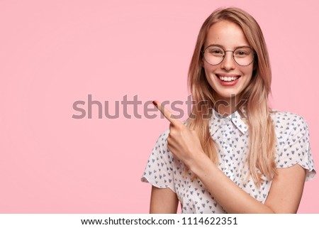 Adorable Caucasian female pupil with friendly look and gentle smile, dressed in fashionable blouse, indicates aside at copy space, shows free place for your promotional text. People and advertisement