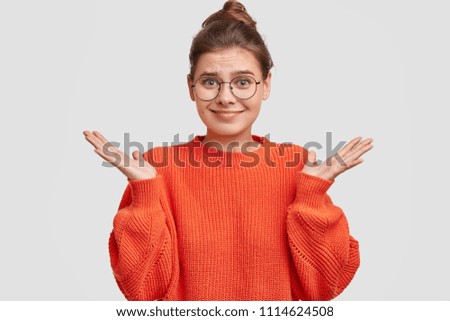 Indifferent pretty young woman shrugs shoulders, wears red sweater, keeps palms raised, poses against white background. Studio shot of adorable female in round spectacles, poses in studio alone Royalty-Free Stock Photo #1114624508