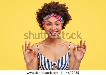 Photo of attractive cheerful satisfied African American female makes okay gesture, smiles gently at camera, isolated over yellow background, wears headband and sailor t shirt, has dark skin.