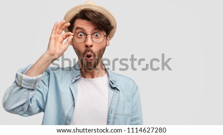 Amazed young European male stares at camera, keeps mouth round, hands on rim of spectacles, wears denim shirt, poses against white concrete wall. People, emotions and facial expressions concept