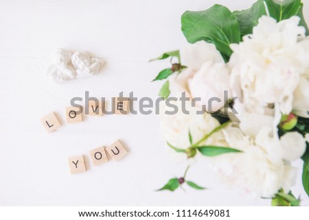 Flat lay Love you lettering spelled in wooden blocks with statuette of two antique lovely angels with blurred white peonies on light background. Love words. Valentine's day. Top view. Copy space