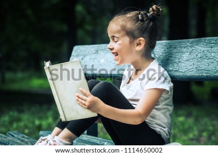 little cute girl is sitting on a bench and is reading a book in the park on a background of green nature