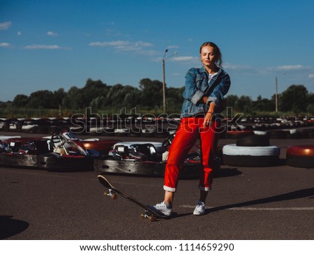 Blonde in sneakers red pants and jeans jacket with skate posing on the asphalt
