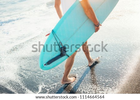 Surfer in white t-shirt walking with surf board near the ocean, legs close-up