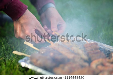 The hand holds a skewer with meat over the coals. Meat is cooked on charcoal on grass. Smoke on meat.