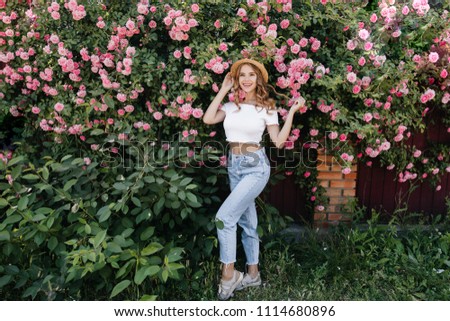Full-length portrait of slim girl in jeans posing near rose bush. Outdoor shot of lovely blonde female model isolated on nature background.