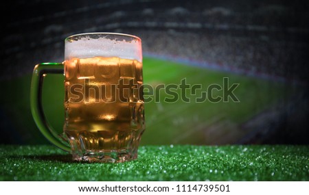 Creative concept. Pint of fresh beer on grass with blurred stadium on background with fog and light. Glass of lager beer ready for drink. Copy space. Selective focus