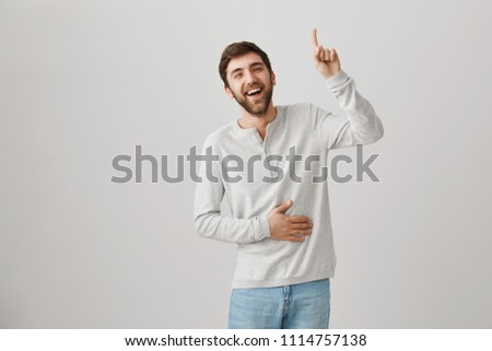 Crazy thought become breakthrough. Studio shot of attractive european boyfriend laughing, holding belly and pointing up with forefinger or showing eureka sign, standing over gray background
