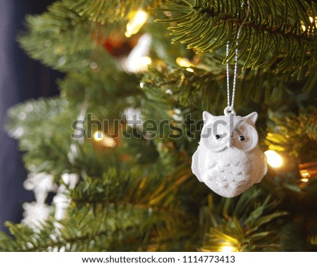 A sparkly white ceramic owl Christmas tree ornament with glitter hangs from a branch of a Christmas tree with glowing golden lights. 