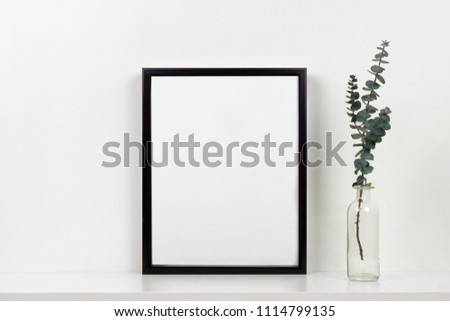 Mock up black frame with vase of branches on a shelf or desk. White shelf and wall. Portrait frame orientation.