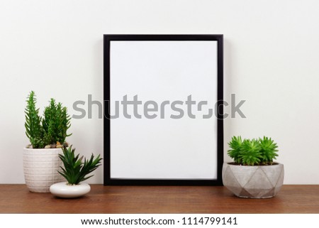 Mock up black frame with succulent plants on a shelf or desk. Wood shelf and white wall. Portrait frame orientation.