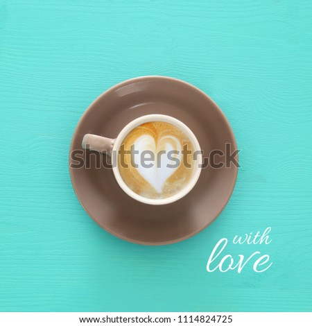 image of coffe cup with foam of heart shape over wooden blue background and text: WITH LOVE