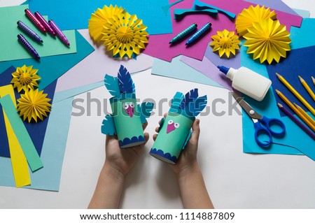 Children's hands make a parrot of colored paper. Toy origami bird. Glue, scissors and paper on a white background. Children's art project for children. Craft favorite.