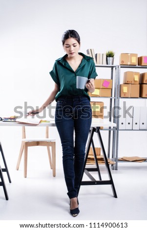 The lady is holdng coffee cup in left hand,hold pen in right hand and put on opened book,thinking about e-commerce,post box for shiping prepare on shelf