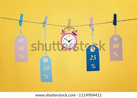 Concept, time for sale. On the rope in the center is a pink alarm clock. Nearby there are labels with inscriptions thirty, fifty and forty percent. Bright photo on a yellow background.