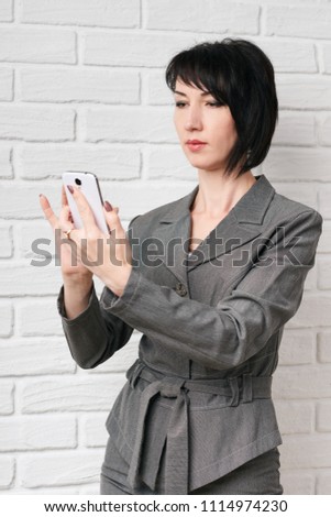 business woman dressed in a gray suit taking selfie on smartphone or preparing to online broadcasting, stay in front of a white wall