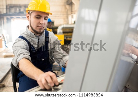 Serious concentrated bearded machine operator in yellow hardhat using control panel while managing conveyor belt at factory workshop