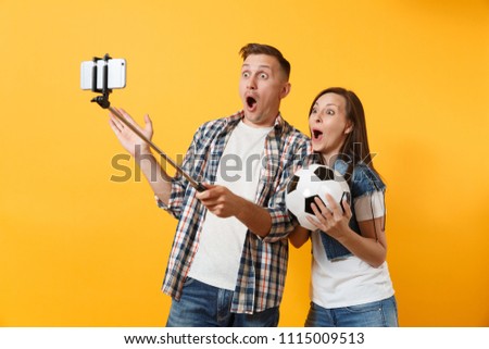 Young couple woman man, football fans doing selfie on mobile phone with monopod selfish stick, cheer up support team, soccer ball isolated on yellow background. Sport family leisure lifestyle concept