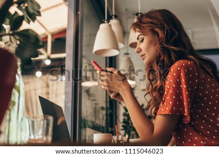 side view of young woman using smartphone at table with laptop in coffee shop Royalty-Free Stock Photo #1115046023