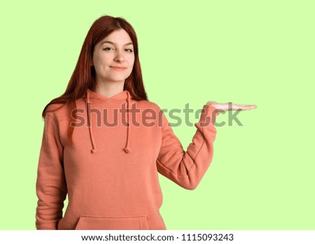 Young redhead girl with pink sweatshirt holding copyspace imaginary on the palm to insert an ad on green background