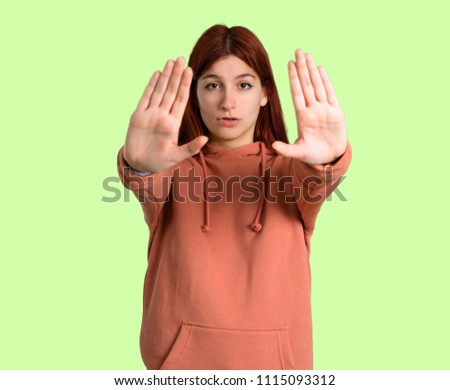 Young redhead girl with pink sweatshirt making stop gesture with her hand for disappointed with an opinion on green background