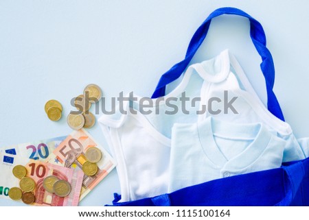 New, light blue baby clothes in the textile shopping bag with euro money on pastel table. Soft colors. 