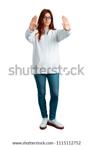 Young redhead girl in an urban white sweatshirt with glasses making stop gesture with her hand denying a situation that thinks wrong on isolated white background