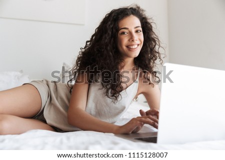 Photo of happy woman with long curly hair wearing silk leisure clothing lying in bed with white clean linen in apartment and using silver laptop