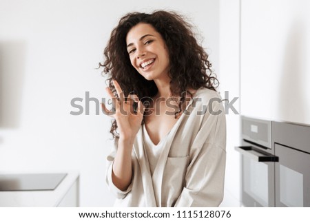 Photo of happy young curly woman with long dark hair wearing silk leisure clothing smiling at camera and showing ok sign while standing in kitchen at home