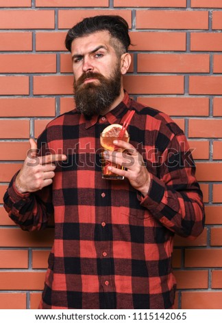 Man points at cocktail on brick wall background. Offer to drink concept. Barman with beard and inquiring face holds glass with cocktail straw. Hipster with stylish hair holds drink or cocktail.