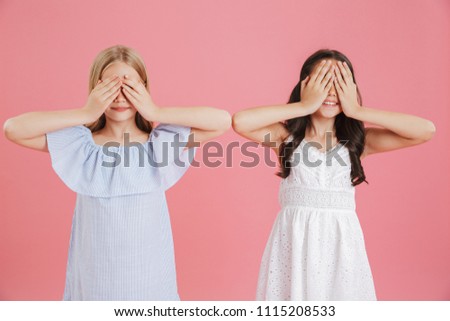 Photo of amusing little girls 8-10 years old wearing dresses having fun and covering their eyes with hands isolated over pink background