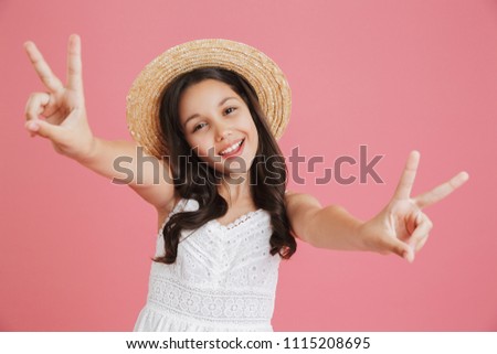 Excited european girl 8-10 wearing white dress and straw hat looking at camera while showing peace sign with both hands isolated over pink background