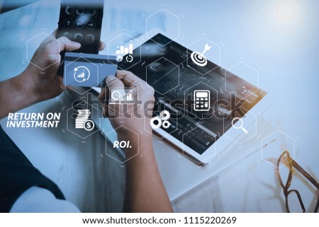 ROI Return on Investment indicator in virtual dashboard for improving business. Businessman hands holding plastic credit card and using digital tablet and laptop computer with smart phone. 