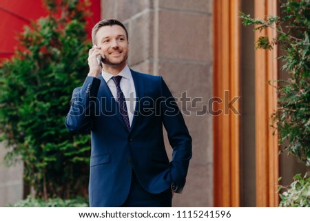 Horizontal shot of glad male entrepreneur speaks via smart phone, looks into distance, wears formal black suit, white shirt and tie, stands outdoor, waits for business partner near cafe or restaurant