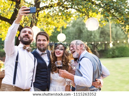 Bride, groom and guests with smartphone taking selfie outside at wedding reception.