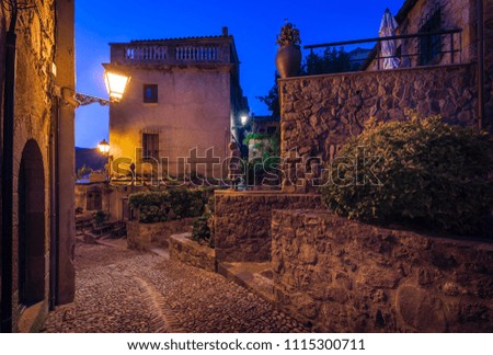 Upper view down on evening street with street lights and houses, girl statue, bush, coarse stone walls and stairs all lit with lamp light under blue dusk sky in ancient town in Costa Brava (Spain)