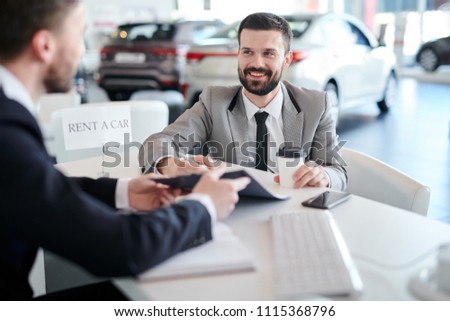 Portrait of mature bearded businessman smiling cheerfully while signing car rental documents sitting across sales manager in car dealership showroom