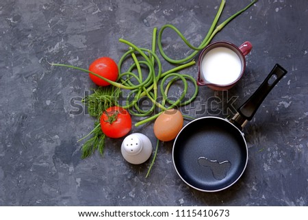 Ingredients for the preparation of an omelette with tomatoes and garlic arrows on a dark gray concrete background. Egg, fresh garlic arrows, tomatoes, milk, dill, salt, frying pan.