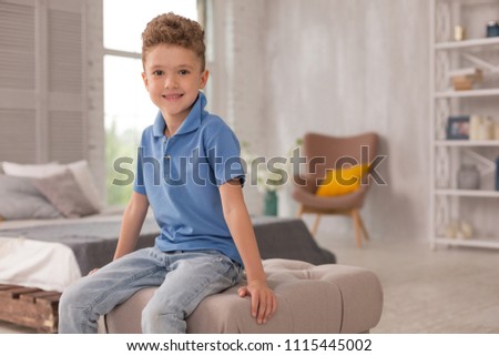 Cute boy. Little pleasant looking cute boy wearing dark blue shirt and denim jeans sitting on hassock in the living room