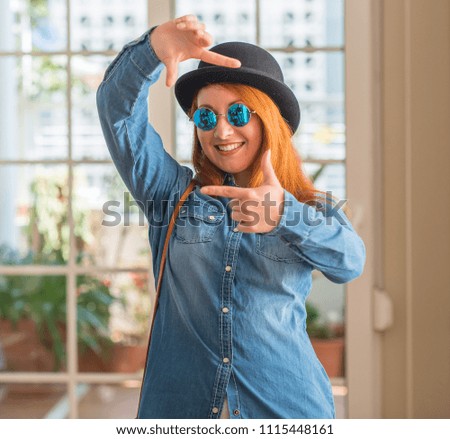 Stylish redhead woman wearing bowler hat and sunglasses smiling making frame with hands and fingers with happy face. Creativity and photography concept.