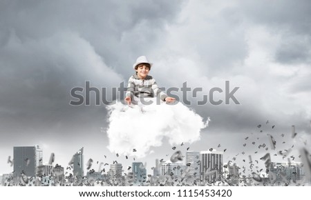 Young little boy keeping eyes closed and looking concentrated while meditating on cloud among flying letters with cityscape view on background.