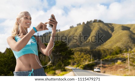 sport, technology and healthy lifestyle concept - smiling young woman with smartphone, earphones and fitness tracker listening to music over big sur hills and road background in california