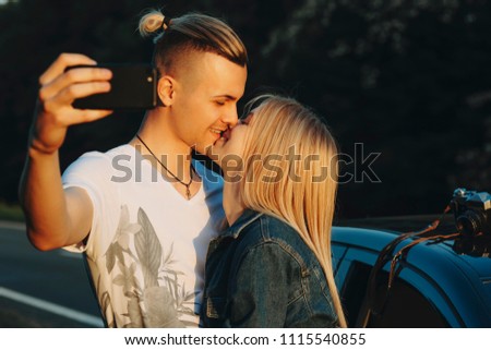 Side view of handsome male kissing attractive blond woman and getting selfie on front camera of smartphone both smiling with closed eyes and standing at car at sunset