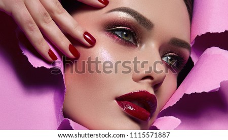 The face of a young beautiful girl with a bright make-up and with plump red lips peeks into a hole in pink paper. Nails with bright red lacquer.