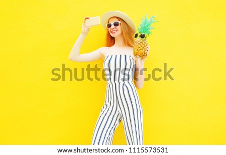 Fashion smiling woman and a pineapple is taking a picture on smartphone in white striped pants, round hat on yellow background