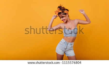 Sporty lovely girl in tank-top and denim shorts dancing with pleasure and smiling. Indoor portrait of stunning female model with vintage hairstyle waving hands on yellow background. Royalty-Free Stock Photo #1115576804