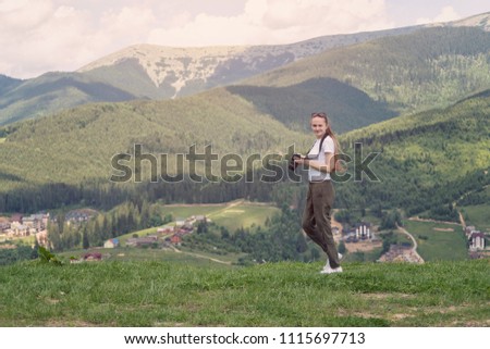 Girl with a displeased face and camera standing on a background of mountains. Forest on background