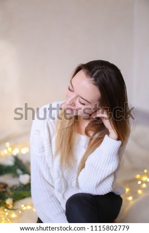 Young nice woman sitting on bed near wreath and yellow garlands. Concept of handmade decorations for Christmas and winter holidays.