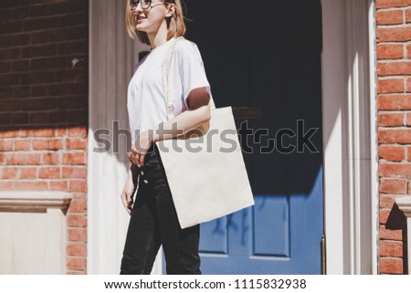 Young hipster smiling girl with white blank cotton shopper paper bag wearing white t-shirt and black jeans, mock-up of blank bag, brick wall and blue door on the background
